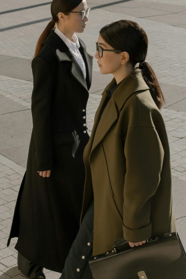 Two businesswomen in trench coats walking in urban area, showcasing modern fashion.